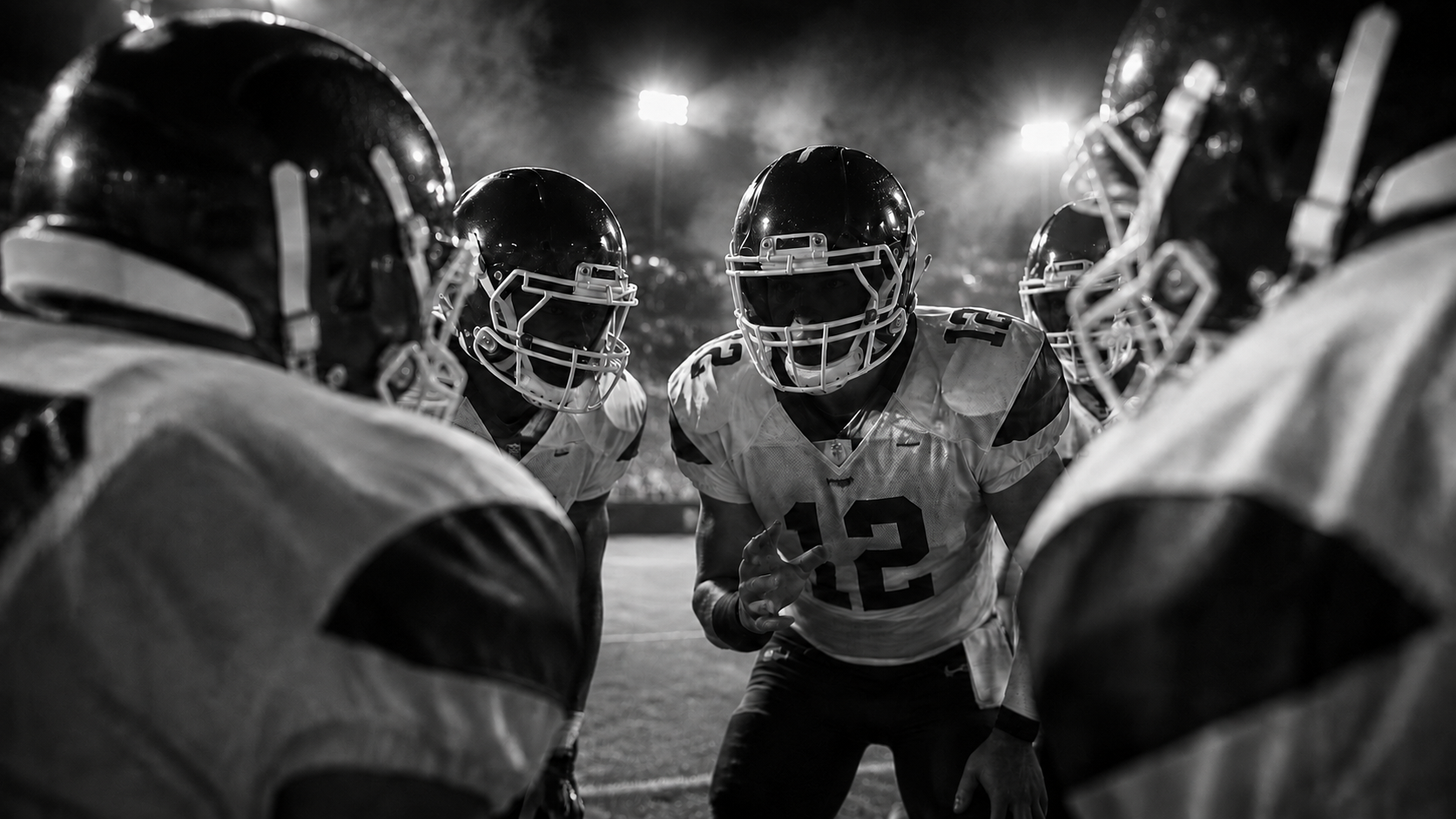 Inside a football huddle, looking over teammates’ shoulders toward the quarterback under stadium lights
